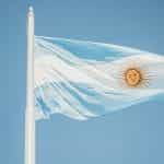 The blue and white Argentine flag waves against a blue sky, on a flagpole.