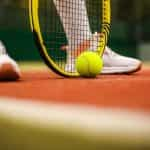 A tennis player’s racket resting on the ground of a clay outdoor tennis court with a tennis ball sitting in front of it.