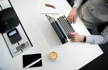 A person using a laptop on a table with a coffee mug, pad, and other gadgets.